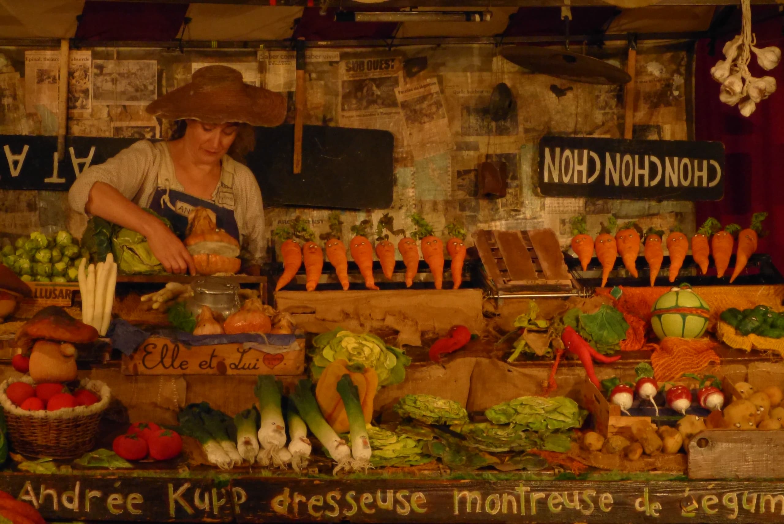 Sur la scène, un artiste manipule un étal de légumes dont les visages sont vivants, dans une mise en scène de marché.