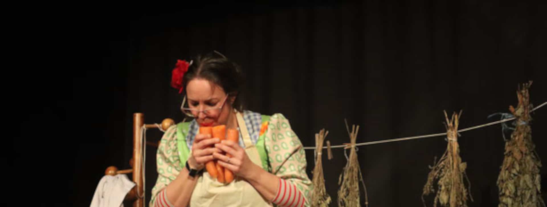 Comédienne qui souffle dans une flûte de pan réalisée avec des carottes, dans un décor de cuisine.