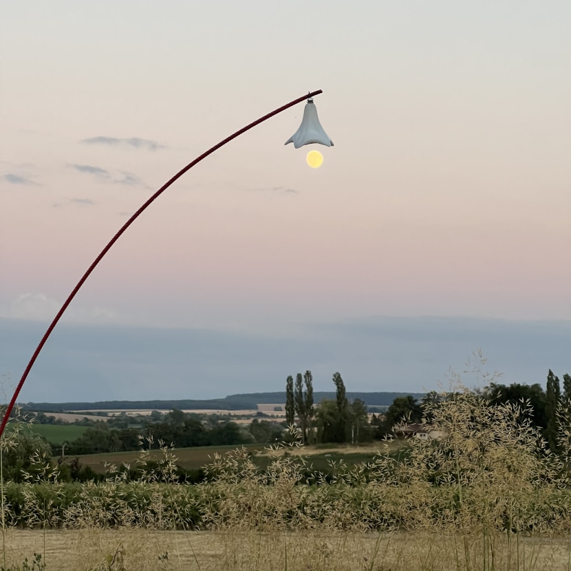 Paysage à la nuit tombante. Un tuteur courbe avec une fleur en verre de Meisenthal suspendue semble faire une cloche à la pleine lune.