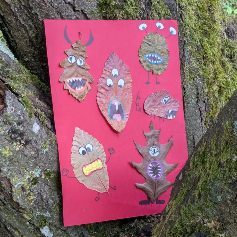 6 monstres grimaçants fabriqués à partir de feuilles mortes, collés sur une feuille rouge, elle même posée dans un creux d'arbre.