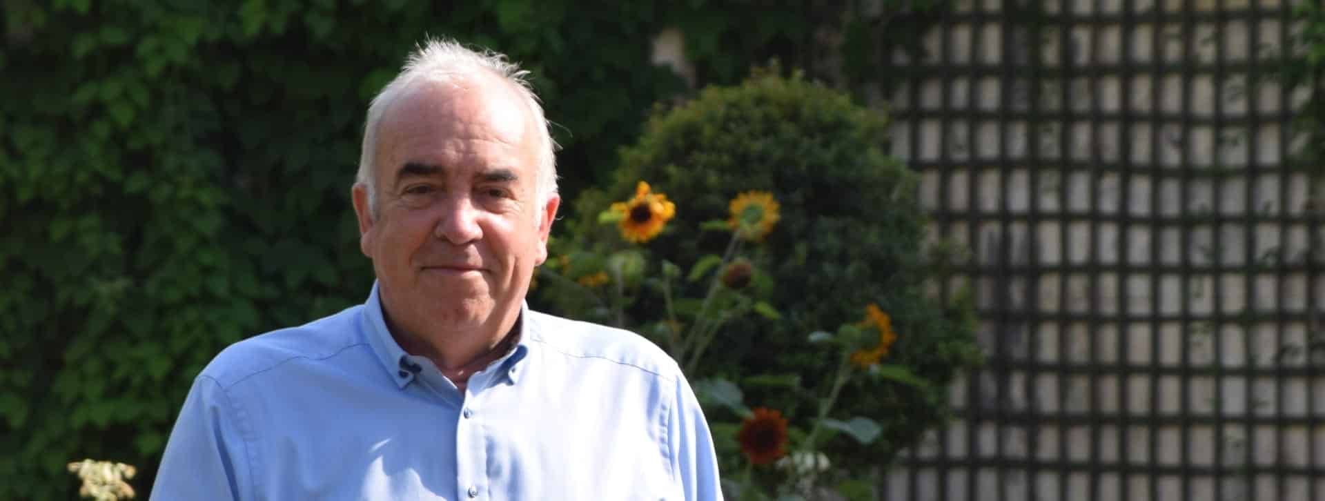 Alain Baraton en bras de chemise, pâquerette à la main, en second plan, un buis taillé en boule et des fleurs de tournesol.