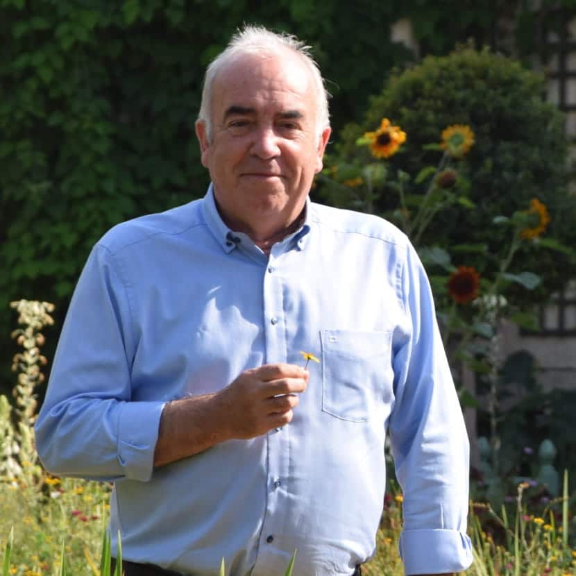 Alain Baraton en bras de chemise, pâquerette à la main, en second plan, un buis taillé en boule et des fleurs de tournesol.