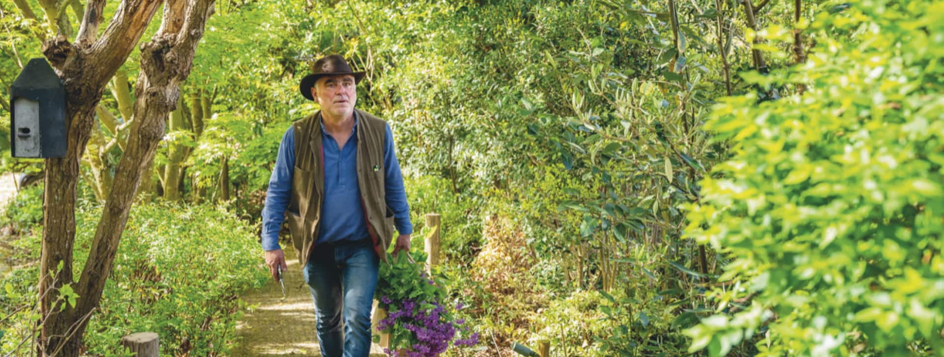 Pierre-Alexandre Risser, chapeau sur la tête, marche sur un platelage en bois avec un bouquet de lilas à la main dans un environnement de sous bois