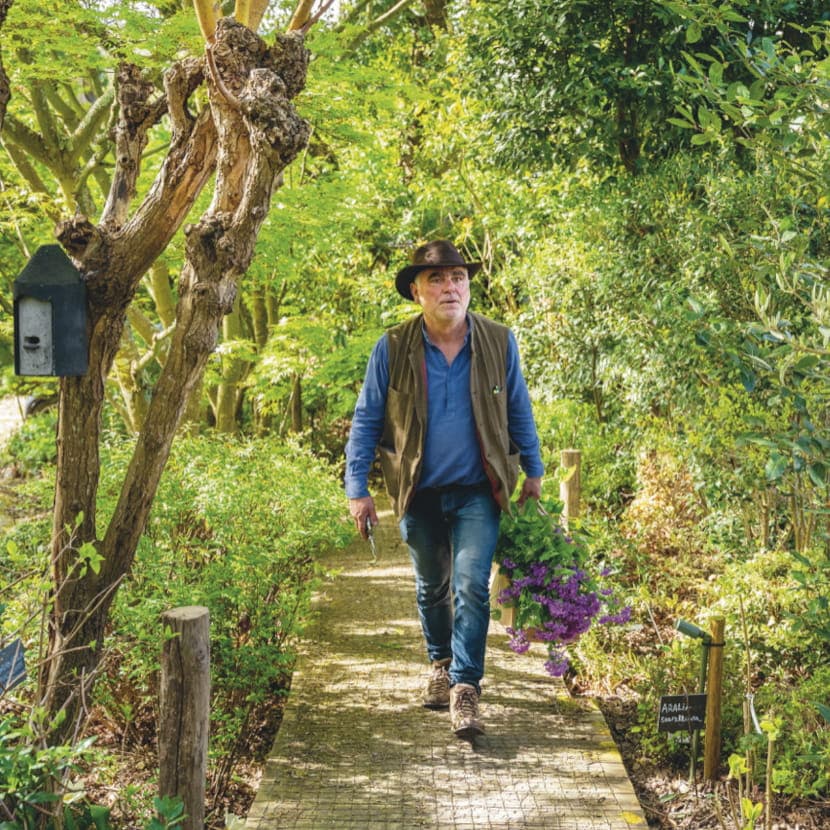 Pierre-Alexandre Risser, chapeau sur la tête, marche sur un platelage en bois avec un bouquet de lilas à la main dans un environnement de sous bois